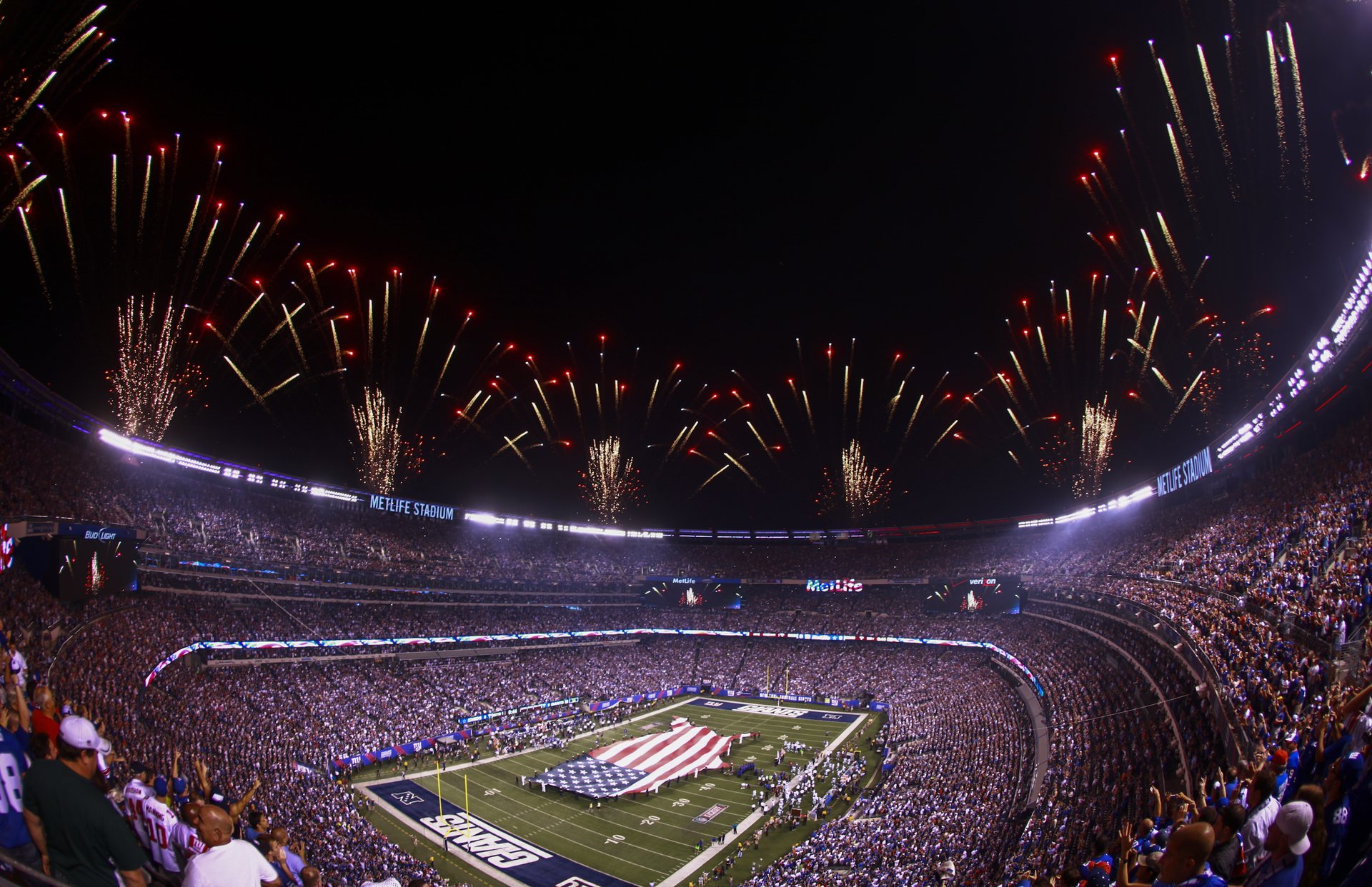 A packed MetLife Stadium at night, with fireworks overhead and a giant American flag unfurled on the field before a Giants game