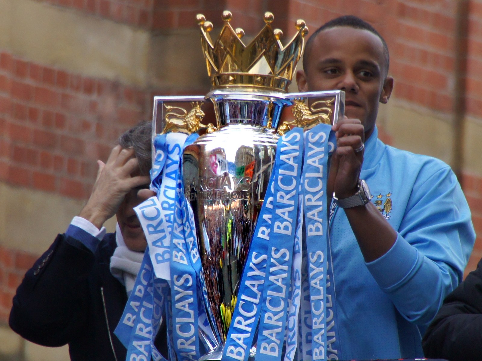 Vincent Kompany holding the Premier League trophy on an open-top bus with Roberto Mancini behind him